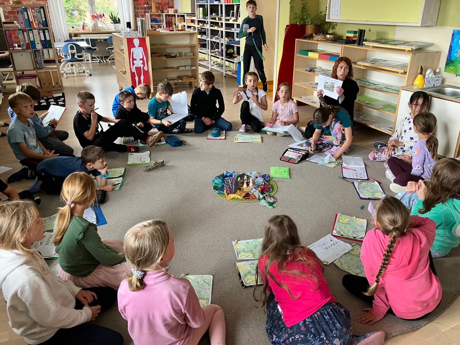 Děti různého věku sedí v kruhu na koberci s pracovními listy.