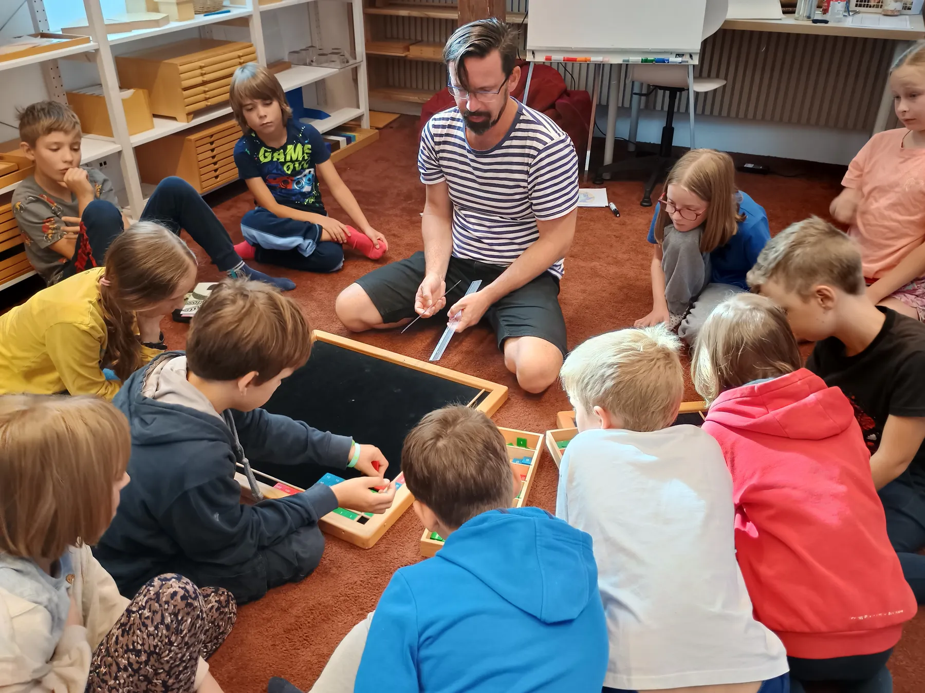 Učitel sedí s dětmi na koberci a společně skládají geometrické tvary z Montessori pomůcek.