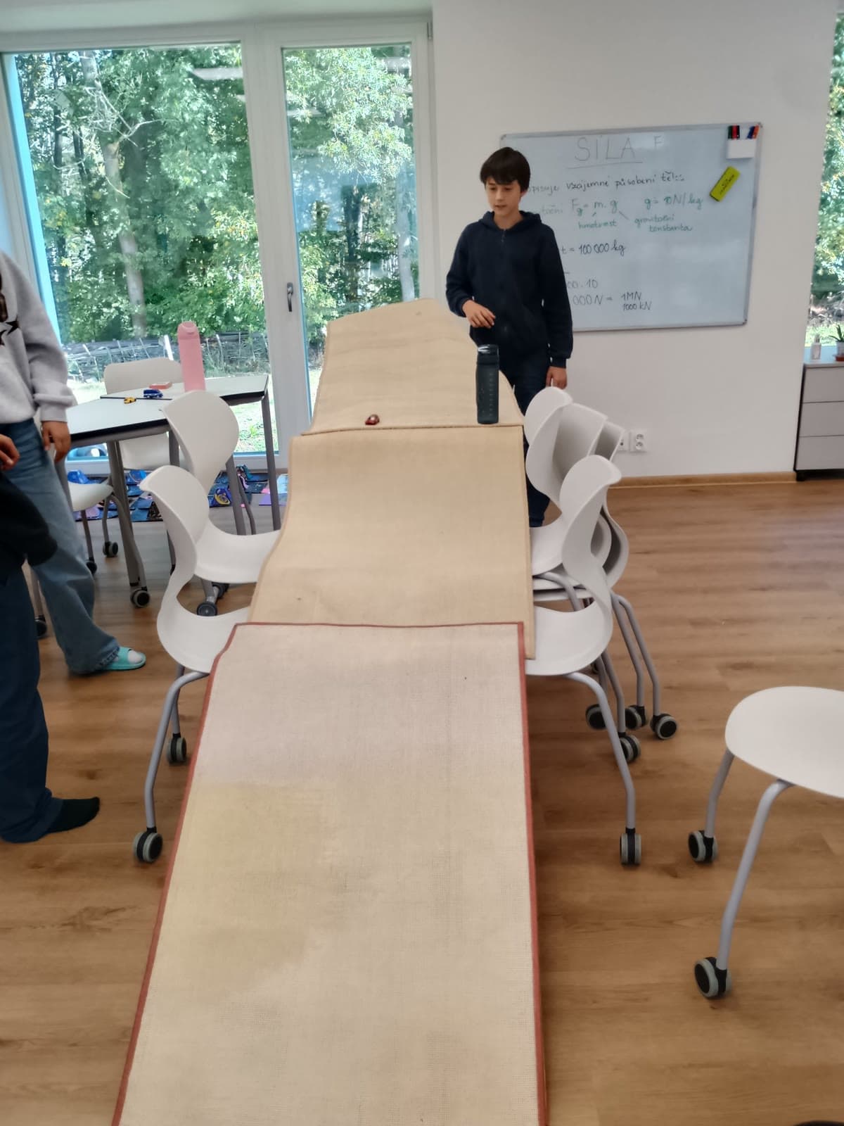 Dráha z koberců vede přes židle z lavice až na podlahu, za ní tabule se vzorcem pro gravitační sílu.