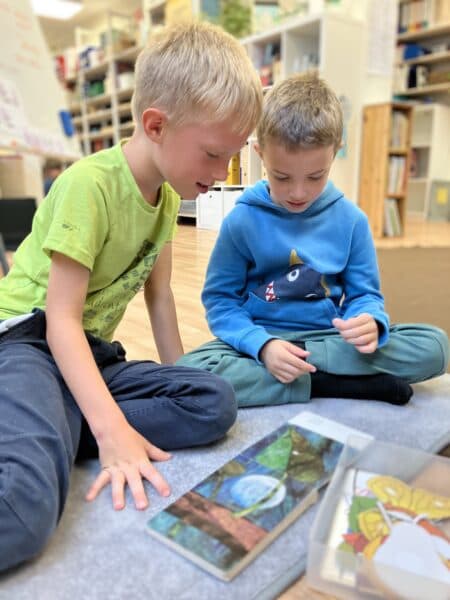 Dva chlapci si spolu prohlížejí pracovní materiál na koberci ve třídě.